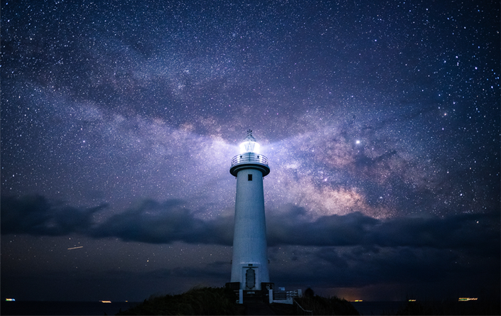 Lighthouse emitting light amid a dark starry, cloudy sky